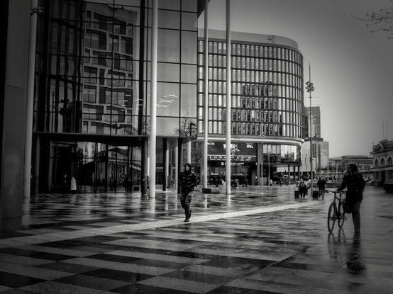 A black and white street photograph of Central Square in Cardiff, captured by ichbinLLOYD. The scene features modern glass-fronted buildings with large columns, reflecting the surrounding architecture on their surfaces. In the foreground, a person walks toward the camera across a checkered, rain-slicked pavement that creates dark reflections. To the right, another person stands with a bicycle. The overall atmosphere is moody and urban, emphasized by a slight vignette around the edges.