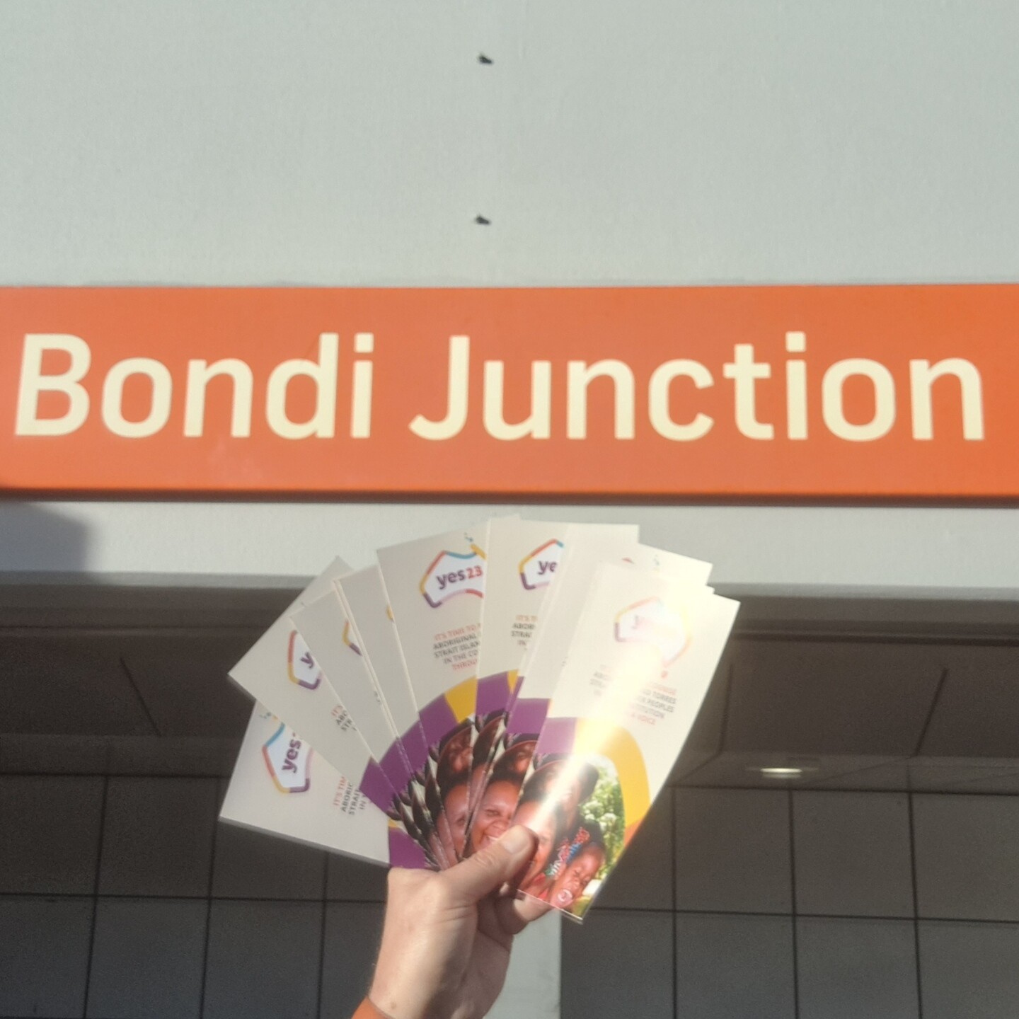 My hand holding Yes23 flyers in front of the Bondi Junction train station sign.