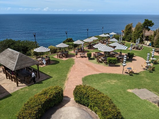 An elevated view of the beer garden on top of the sea cliff with grass, outdoor tables and blue/white striped umbrella. In the background is the sky and the Pacific Ocean.