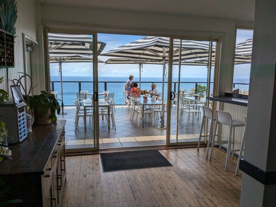 A view from the bar through to the upper dining terrace with tables and blue/white striped umbrellas. In the background is the sky and the Pacific Ocean.