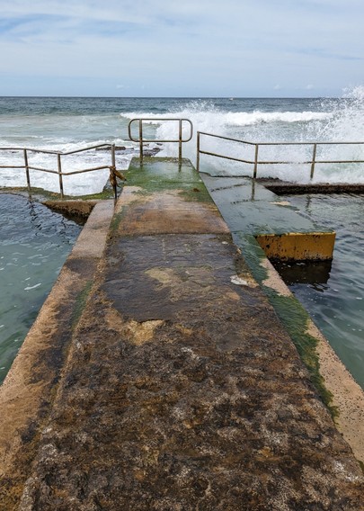 The end of the concrete walkway between the two pools. Beyond, waves crash and spray against the rocks. The walkway has a thin bed of seaweed. Small whisps of seaweed can be seed clinging to the stainless steel handrail at the end of the pools.