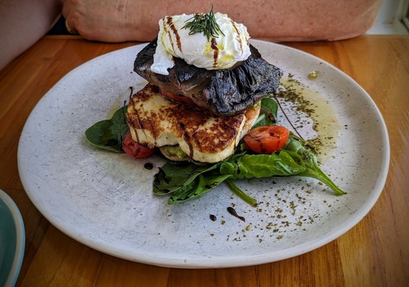 A grey plate sits on a wooden table. On it is half an English muffin, surrounded by spinach leaves and halved tomatoes. On the muffin is fried haloumi, fried mushroom, avocado and a poached egg, all drizzled with olive oil and balsamic glaze.