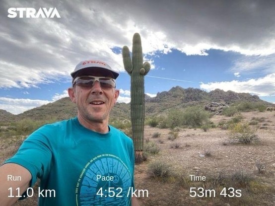 A man taking a selfie with a desert landscape in the background, featuring a large saguaro cactus and mountainous terrain. The overlay text shows running statistics from the Strava app, indicating a run of 11.0 km, 4:52/km