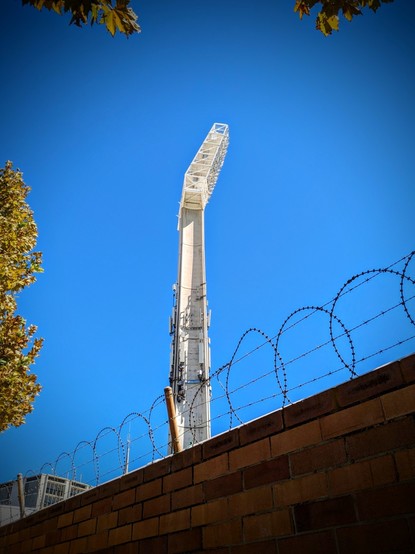 A concrete light pylon in full sun, behind a red brick wall topped by razor wire. There are various cables and UHF antennas around the pylon. Tree foliage fringes the image against a cloudless blue sky.