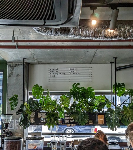 The inside of the cafe with the chrome espresso machine at the bottom of frame, several monstera plants covering the windows, various pipes and ducts hanging from the bare concrete ceiling. Above the windows is the menu that simply reads:
"
BLACK       CHOC
WHITE       CHAI
BATCH BREW        MATCHA
TEA
"