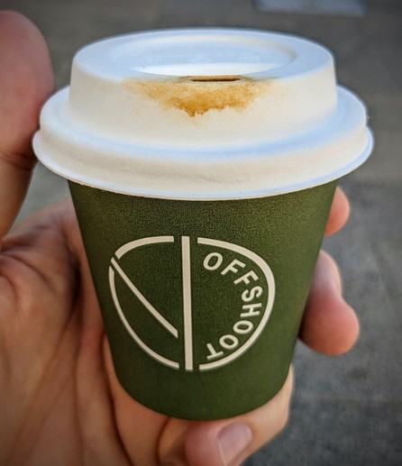 A hand holding a small, olive coloured paper coffee cup bearing the "offshoot" logo. The white paper lid has blotchy brown staining around the sipping hole.