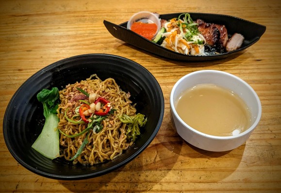 The dishes: a large, round, black bowl containing fried mee with some vegetable garnish; a boat-shaped, black dish with sliced char siu, a cabbage salad and a small cup of chili sauce; a small, white, round bowl of tan coloured broth.