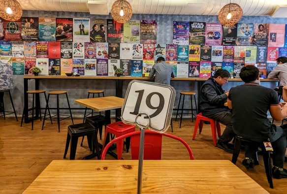 A view across the restaurant with small posters adorning the far wall. There are several diners in the middle distance and on the table in the foreground is a standing service ticket with number 19 on it.