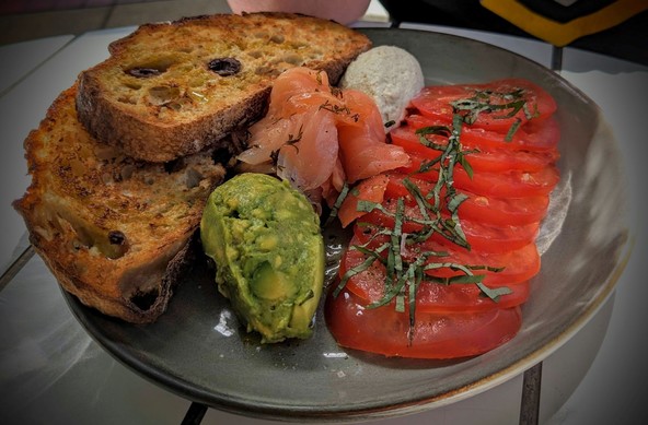 The Healthy Start - On the right side of the plate are slices of ripe tomato with a chiffonade of basil over them.  On the left are two slices of toasted olive sourdough bread. In the centre of the plate is a mound of smoked salmon. Above and below the salmon are quenelles of cashew/cauliflower cheese and smashed avocado.