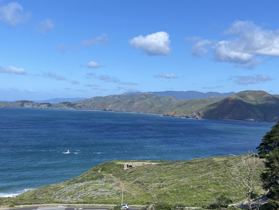 A scenic coastline with blue ocean waters, lush green hills, and a clear sky with scattered clouds. A road is visible at the bottom of the image.