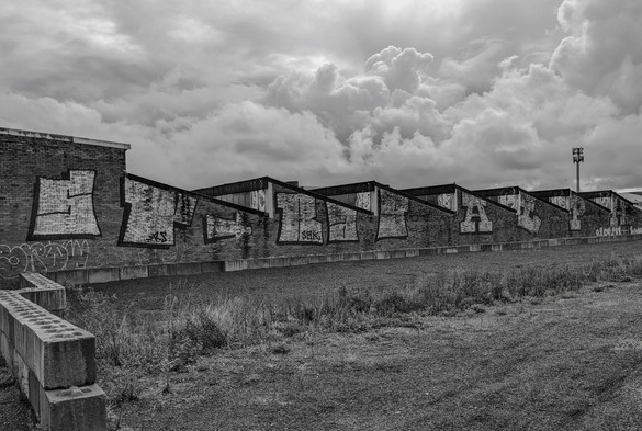 A black and white photo of the side of some "sawtooth roof" warehouse buildings with graffiti on them. Dramatic clouds billow in the background.