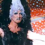 A GIF of actor Terrence Stamp, an older man with pale skin, dressed in heavy make-up and wearing an emu costume in the film Priscilla Queen of the Desert. He is dancing, grudgingly, in front of Indigenous Australian art.