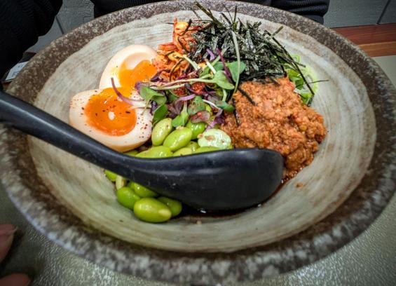 A bowl of spicy pork mazesoba. A soy boiled egg, edamame beans, ground pork, micro herbs and a chiffonade of dried seaweed is visible on top.