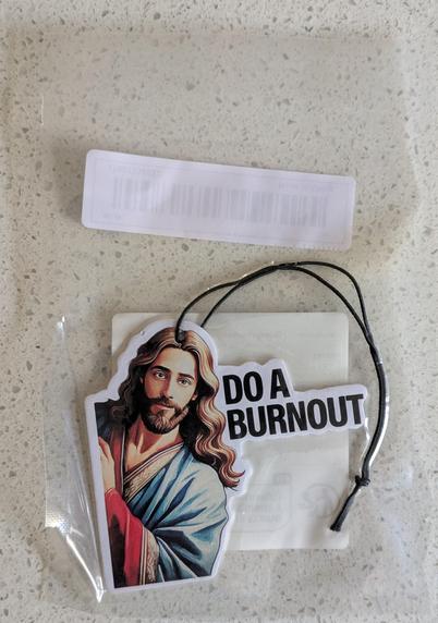 A cardboard hanging deodoriser inside a sealed plastic bag. It features an image of Jesus learning in, saying "DO A BURNOUT".