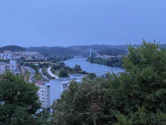 A river viewed from above at dusk. A bridge across a river is visible in the distance with rooftops in the foreground 
