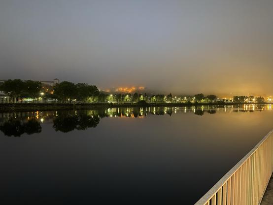 A wide river in fog near dawn with lights visible in nearby buildings 