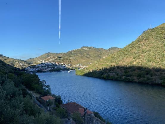 A scenic view of a river surrounded by green hills, with a small town on the hillside. The sky is clear blue, and there is a faint airplane contrail visible in the upper part of the image.