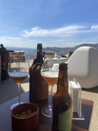 A pair of beer bottles and glasses of beer with a backdrop of mountains and blue sky.