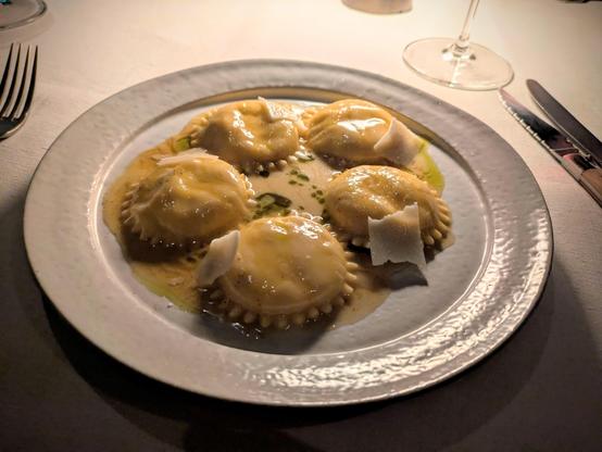A white plate with 5 round ravioli with crimped edges covered in a beige sauce with a few drops of green oil floating on it and a couple of shavings of parmesan.