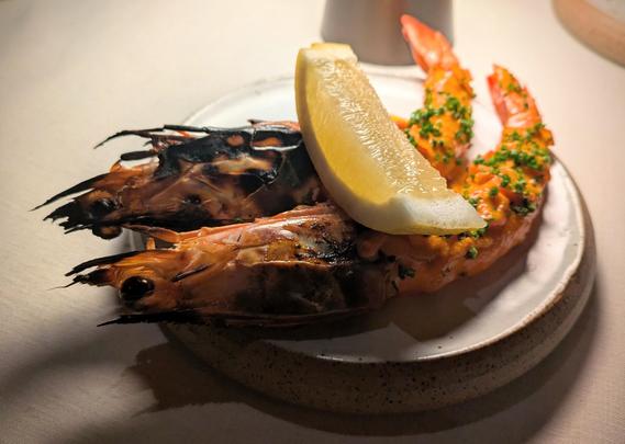 Two charred prawns sitting on a plate. The prawns heads and tails are intact, but their intervening shells have been removed and covered in an orangey-red sauce with a sprinkling of finely chopped chives. Sitting on top is a wedge of lemon.