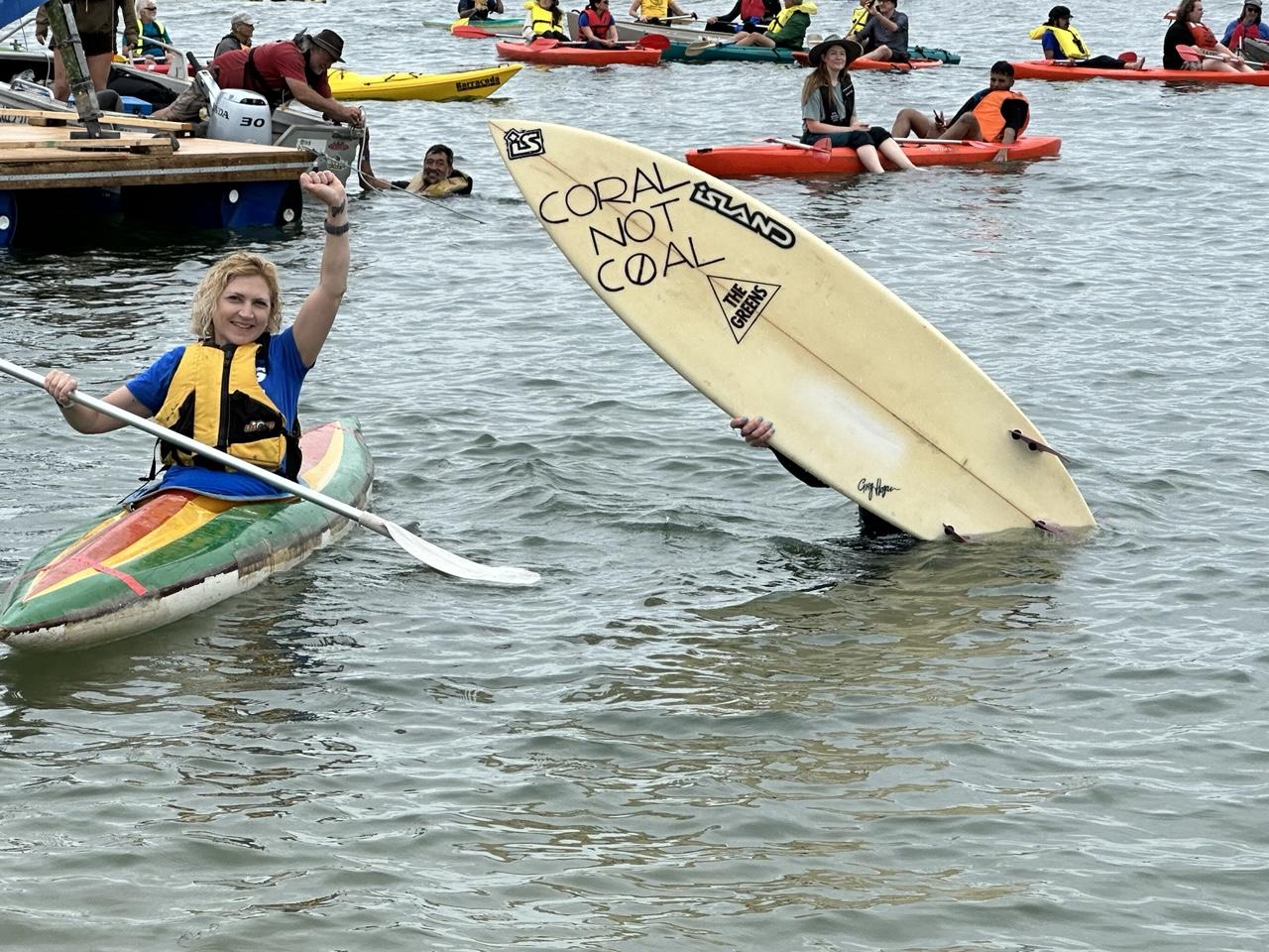 Woman in rainbow-painted kayak with one fist raised, next to person holding a surfboard in front of them with “Coral Not Coal” written on it. Lots of other kayaks on the water in background.