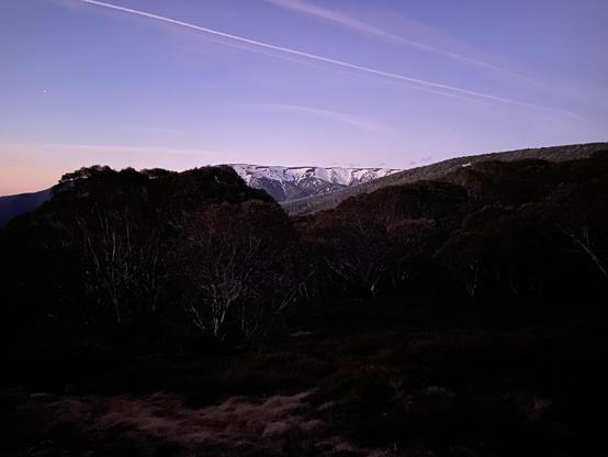 A snow capped mountain at dusk viewed across a dark valley under purple skies with contrails visible.