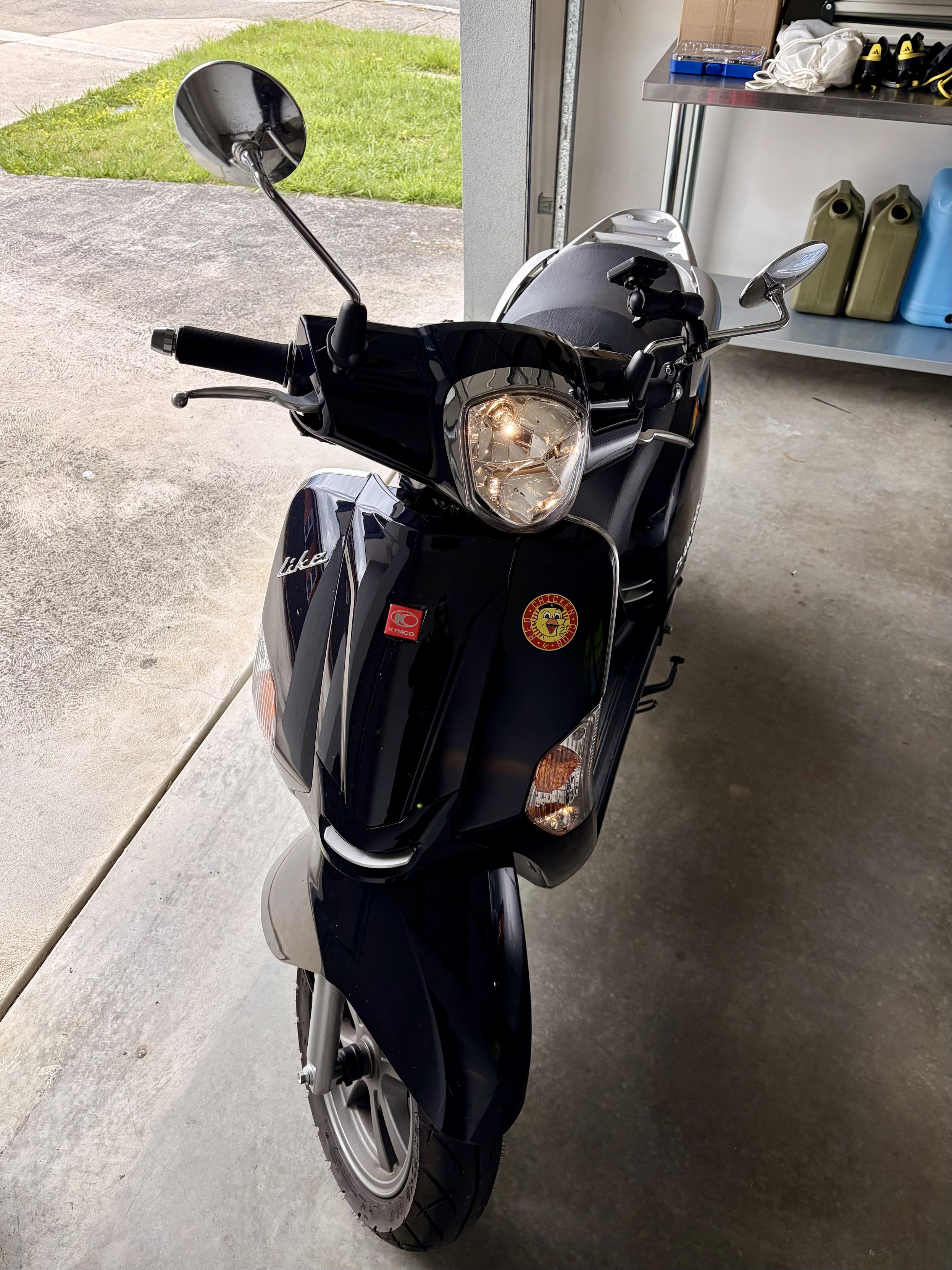 A black scooter in the doorway of a garage. It is just insdie and the photo is from the front. The lights are on. You can see a driveway and grass outside and a bench is behind it. The scooter is pretty barebones now.