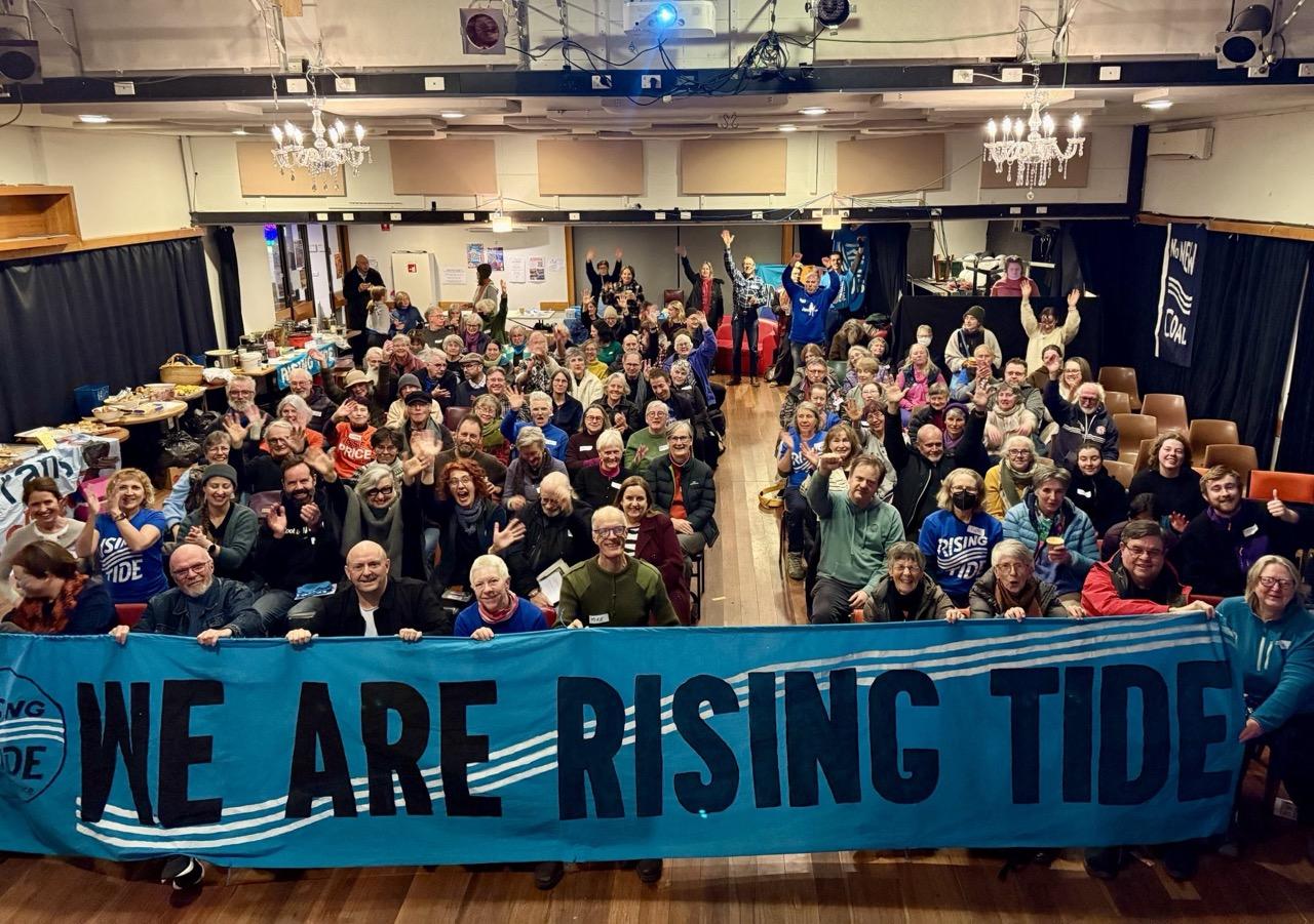 Hall full of happy people holding a big blue banner with “We Are Rising Tide” on it