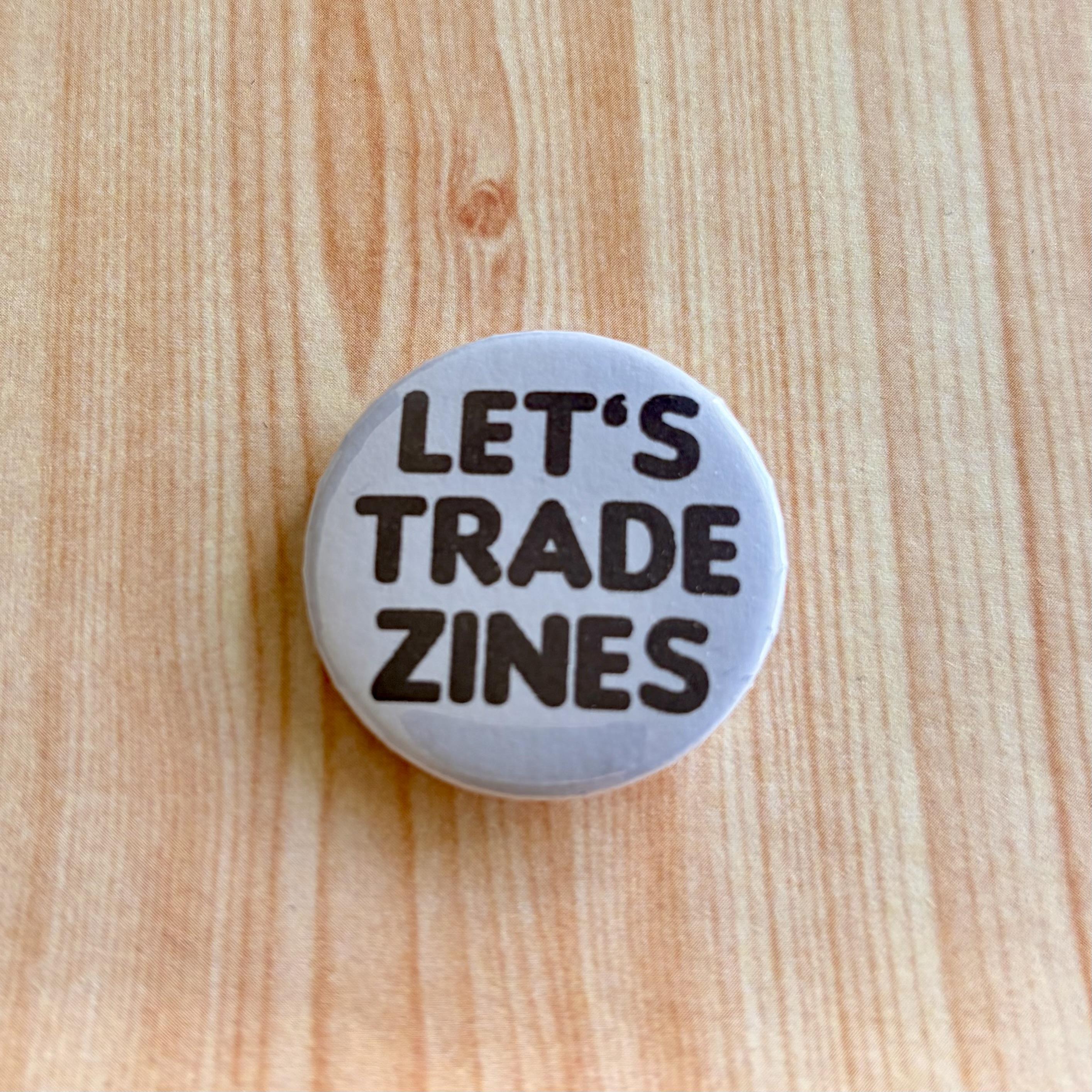 A little round badge that says “Let’s Trade Zines”.  White with black block text. It sits on a piece of timber paper.