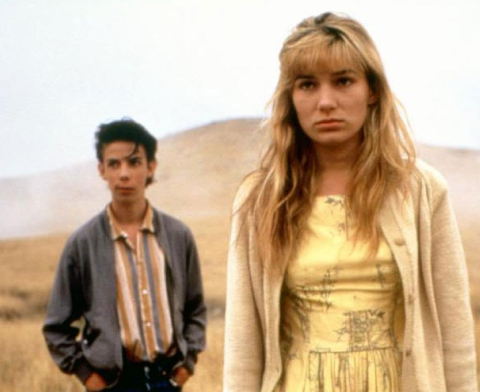 A screen shot from the movie. There is teen aged girl in a peach cardigan and yellow dress. She has sandy hair and a frown. Behind is a young man, very skinny and very young. in a button up shirt and a bomber jacket, he is also grumpy. They are in the desert.