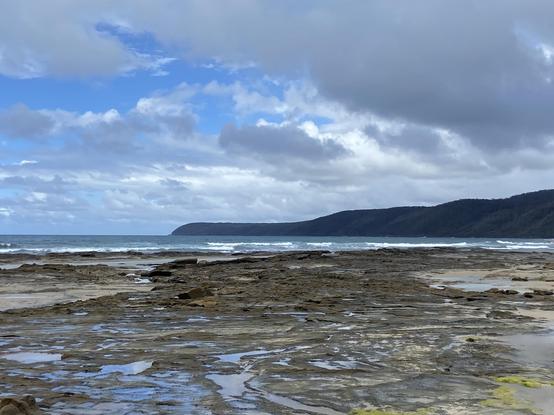 A view across a beach with hills visible in the distance under partly cloudy skies.