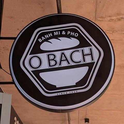 A round white-on black shop sign with a hexagonal logo for the "O BACH" restaurant. The top of the logo has the text "BANH MI & PHO" with the image of a bread roll. The lower side of the logo has the image of a bowl with the text "SINCE 2020".