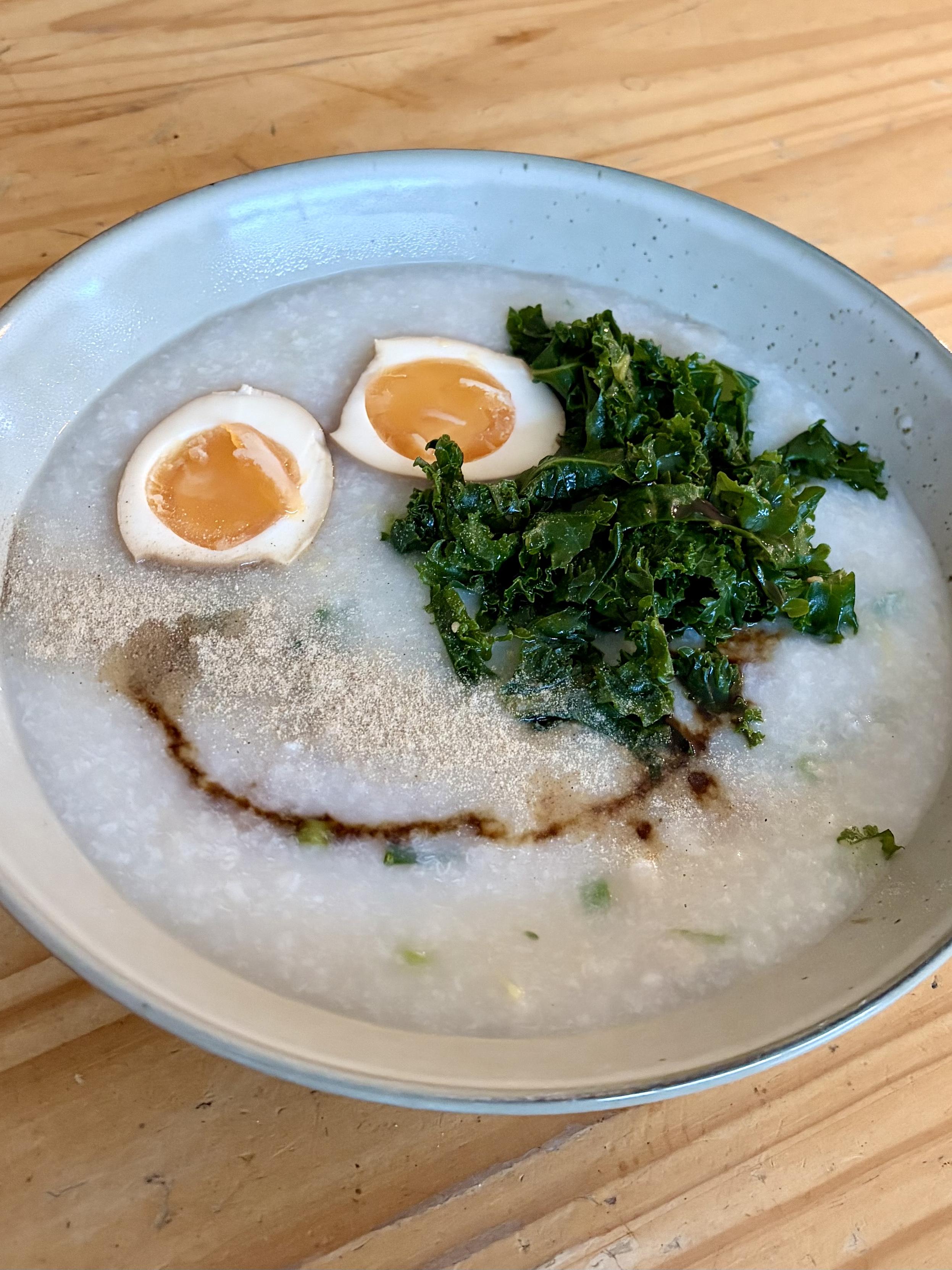 A bowl of white porridge with two halves of an egg at the top. A bit of kale on one side and a mouthy smile made with a line of soy sauce.