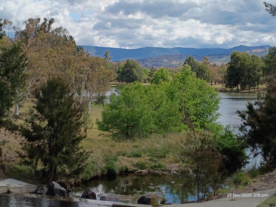 Trees of varying shades of green, a catchment pond, a bluish lake, dark blue mountains, and above it all a blue sky filled with fluffy white clouds. 