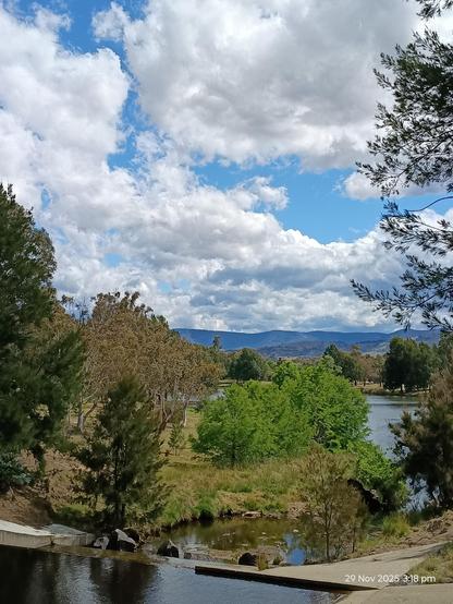Trees of varying shades of green, a bluish lake, dark blue mountains, and above it all a blue sky filled with fluffy white clouds. 