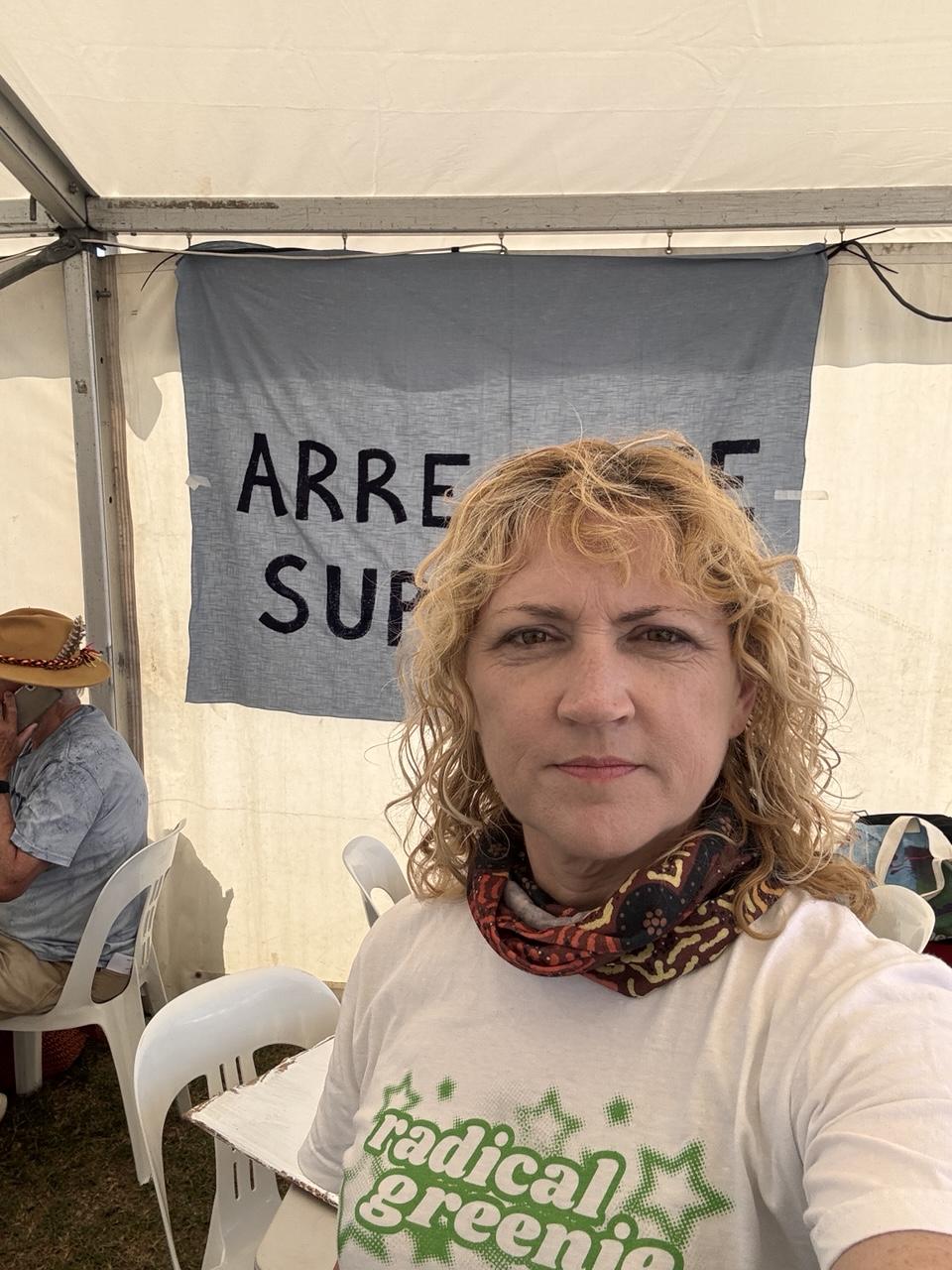 Emma in a Radical Greenie shirt and neck scarf standing in front of blue Arrestee Support sign in a white marquee