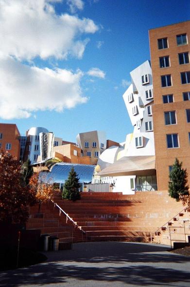 A small ampitheatre in front of a cluster of modern buildings of various shapes, some brick, some reflective, under a blue sky with white clouds. A few small trees are planted around the ampitheatre