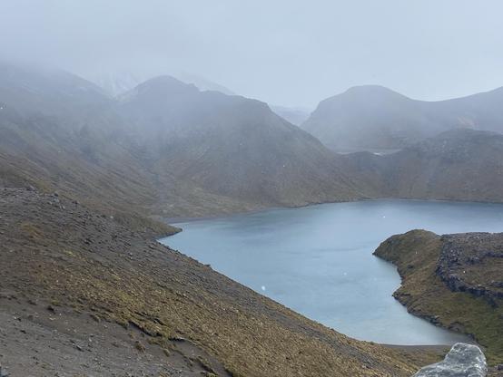 A volcanic lake shrouded in mist, surrounded by mountains. Light snow is falling and snow capped peaks can be seen in the distance.