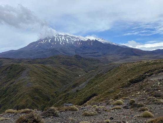 A large and rugged mountain with patches of snow under partly cloudy skies with cloud touching the top of the peak. Treeless scrub covers the slopes in the foreground.