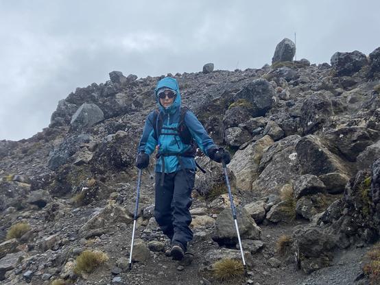 A woman dressed head to toe in waterproof gear hikes down a rocky slope and balances with hiking poles.