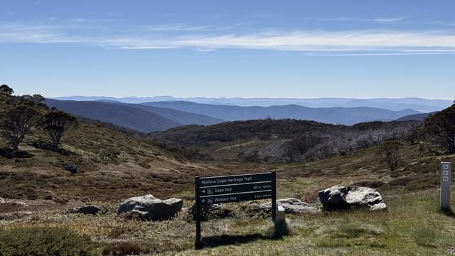 Rows of mountains shrouded in blue mist under a bright blue sky. In the foreground is low grass, some rocks, and a sign “Wallace Heritage trail” pointing to Wallace Hut and Cope Hut.