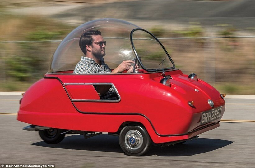 A hipster-looking man in a checked shirt and sunglasses, driving an incredibly tiny red, three wheeled microcar (the Peel Trident)