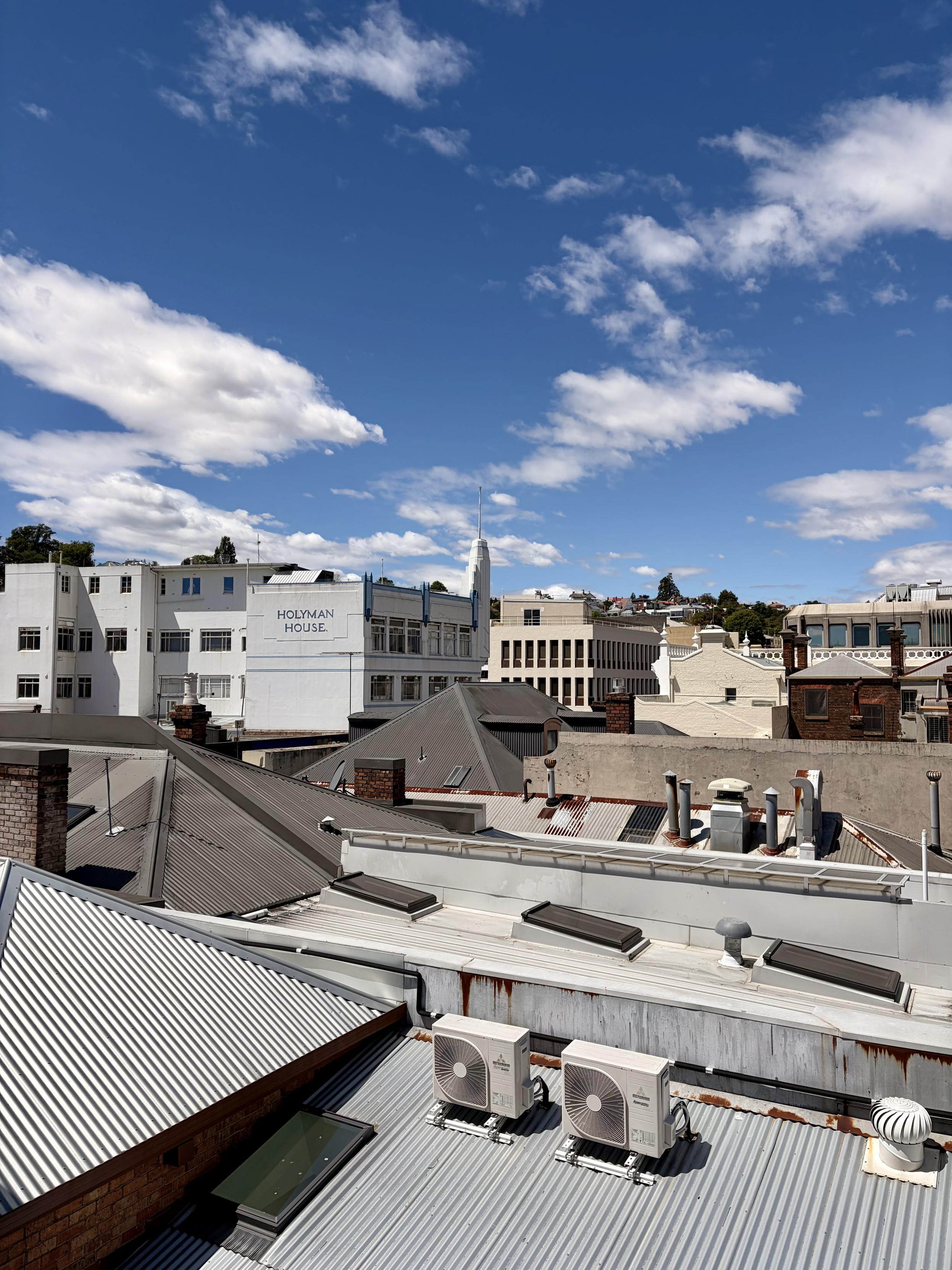 A deep blue sky with fluffy clouds above rooftops with air conditioning units, tin roofs and art deco buildings. Kind of industrial.