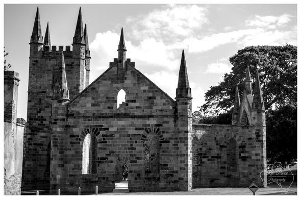 A dramatic black and white photograph of the roofless ruins of the Port Arthur convict church. The image highlights the sharp, pointed spires and intricate stonework against a bright, cloudy sky, capturing the haunting Gothic architecture of the site.