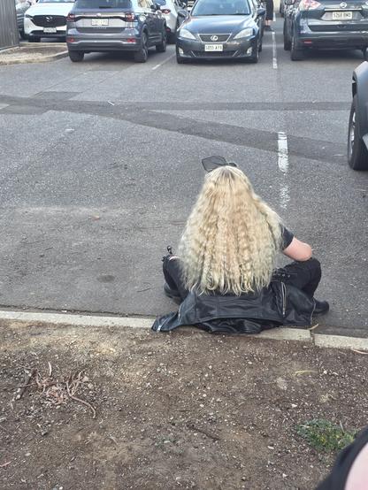 Back of a girl with crimped blonde hair in black jeans and a faux leather jacket sitting on the curb in a carpark