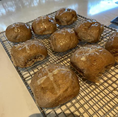 Nine hot cross buns on a cooling rack with light reflecting off the glaze. Some have crosses visible, some not so much. 