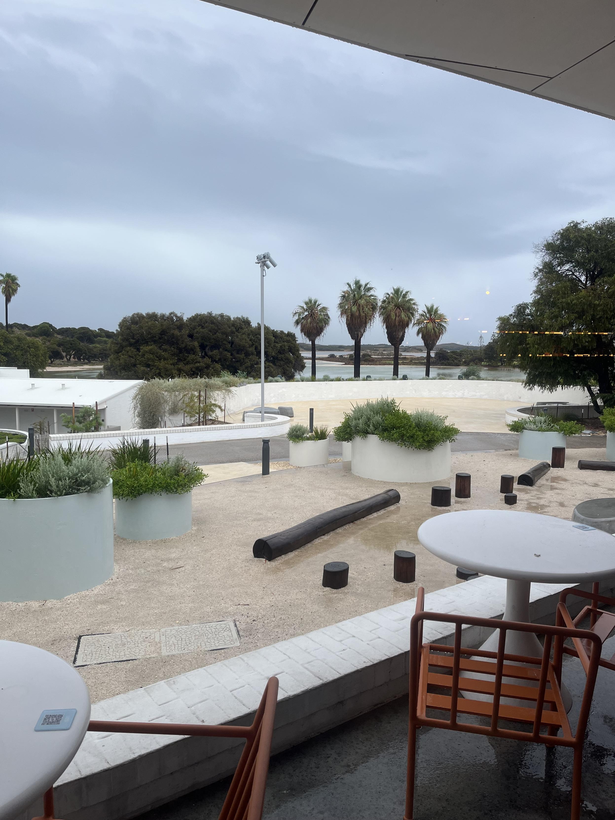 A photo from a bar looking out across a wet sandy terrace with palm trees and a salt lake in the distance. Grey clouds mottle the sky. Sunsets at the Settlement on Rottnest Island