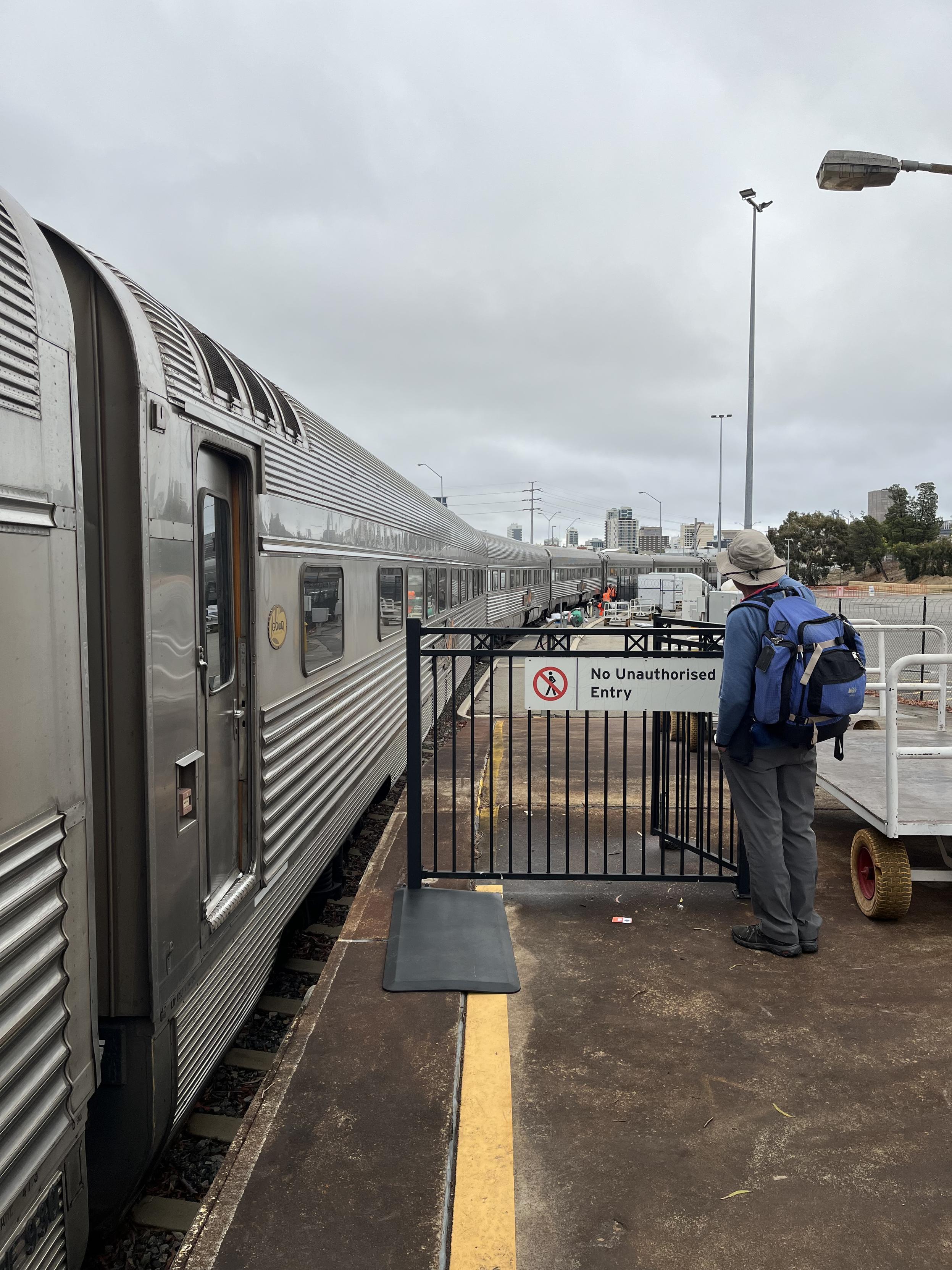 A photo of the end of a train platform with a fence blocking passengers from walking further. A man stands looking at a train as it extends into the distance. Workers in orange vests load things onto the train. Waiting to board the Indian Pacific