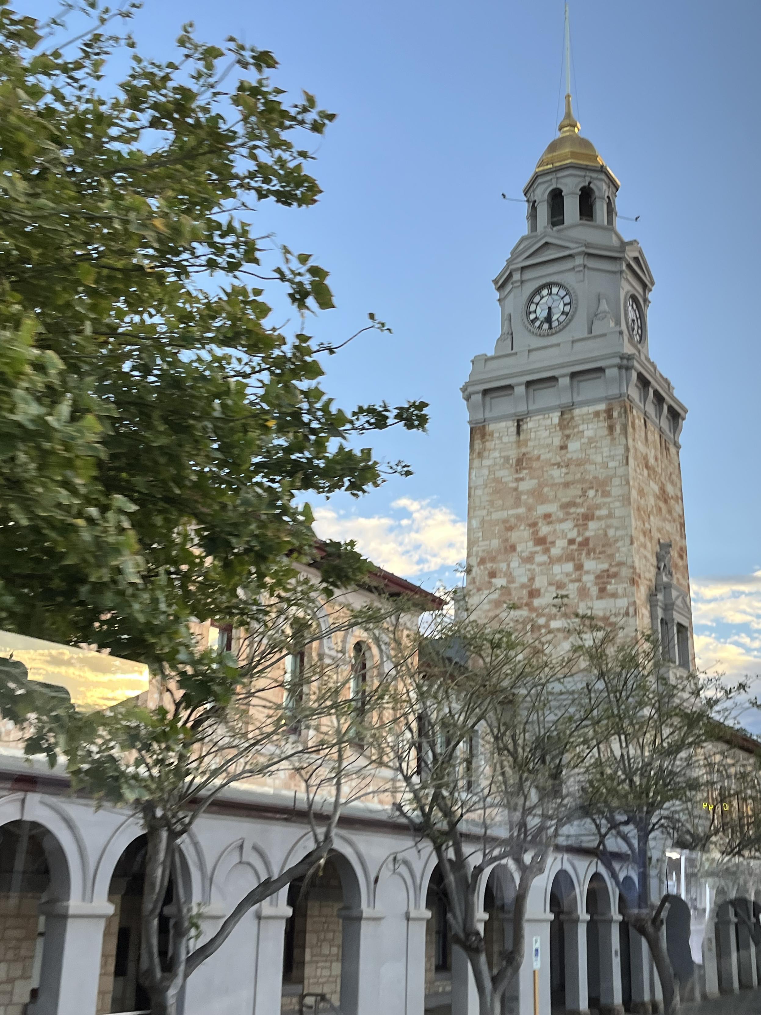 A photo of the clock tower in Kalgoorlie which is topped with gold leaf. A tree frames the shot on the left and a bright blue sky backs the view. The gold leaf clock tower of Kalgoorlie