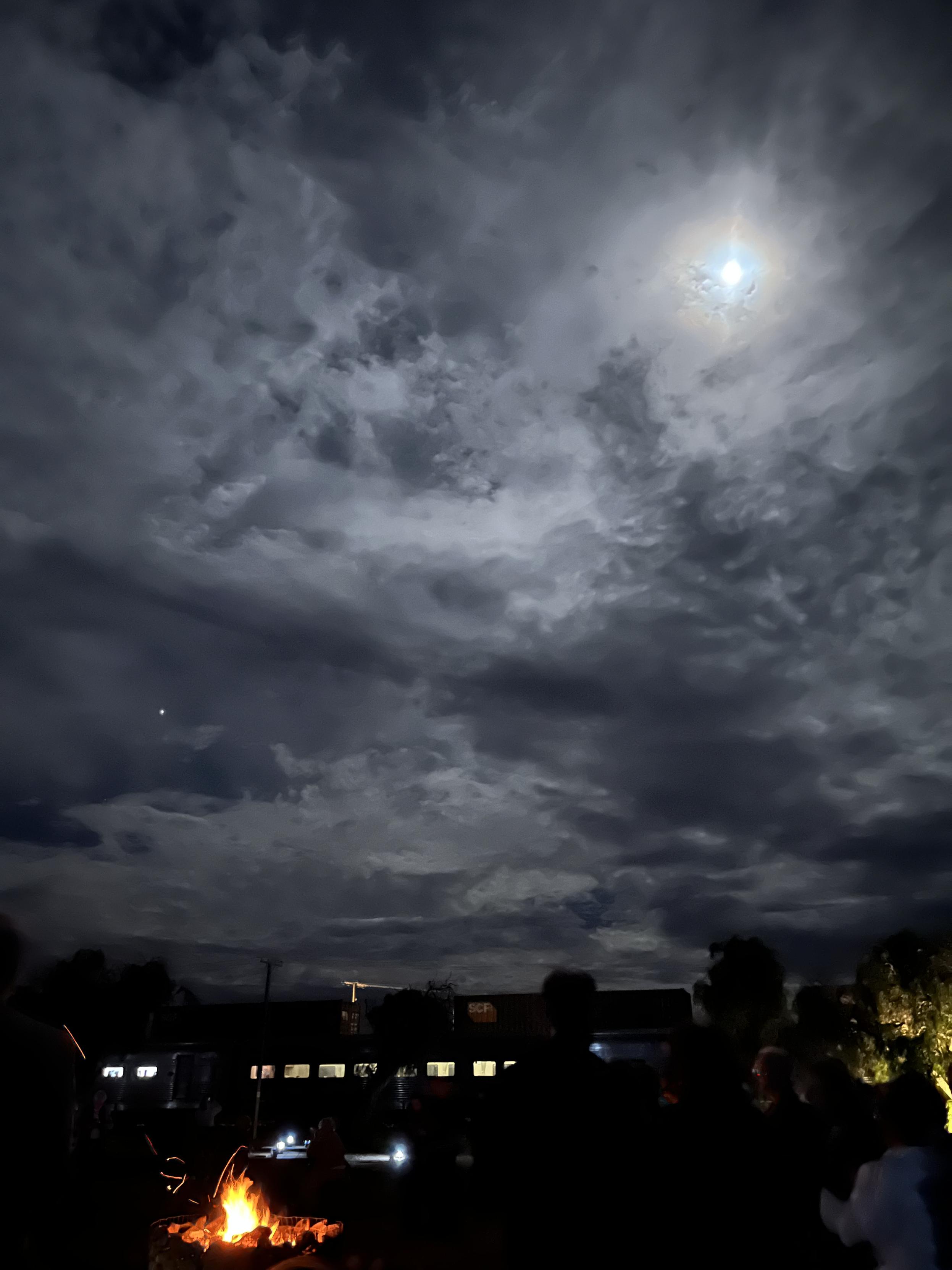 A photo of the night sky in Cook, WA. High thin clouds cover most of the stars, but a gibbous moon glows brightly through. On the ground below, people stand around a bonfire, and a train waits in the background with windows aglow. Bonfire and moonlight at Cook